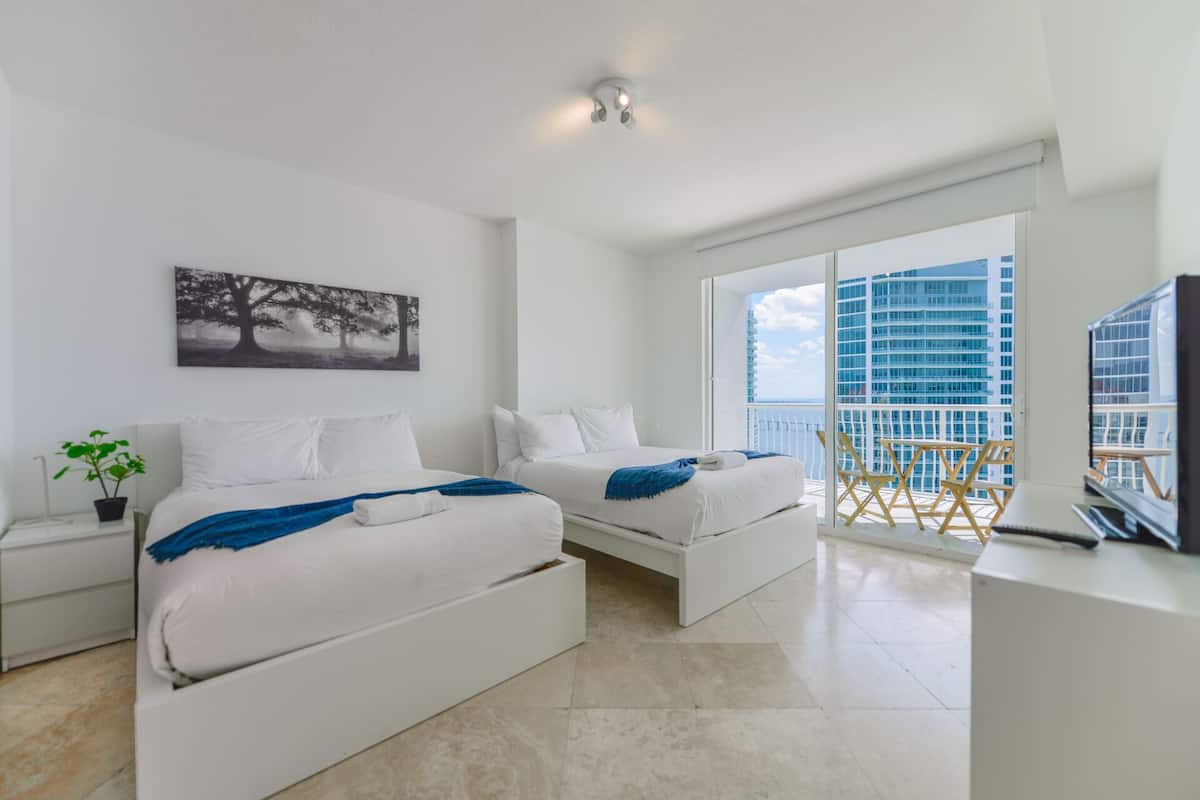 A modern hotel room with two beds, a balcony with a view of a high-rise building, and a mounted artwork on the wall.