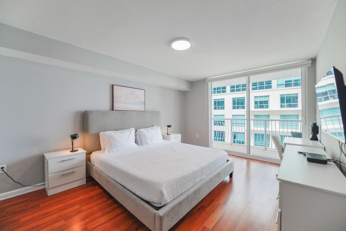 A modern bedroom with a large bed, bedside tables, a desk, and a window with a city view.