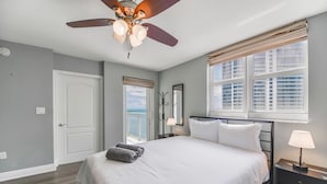 Un dormitorio con cama, ventilador de techo, ventana con persianas y vista al océano.