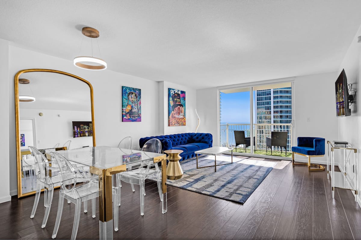A modern living room with a clear glass dining table, a blue sofa, and a large mirror.