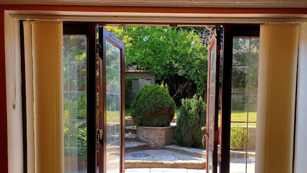 A room with a sliding glass door leading to a patio area with a stone floor, a small garden, and a view of a tree-lined street. - Lovely 4-bed , 3 Bath Villa in Bolton, Manchester (Bolton)