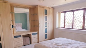 A bedroom with a bed, a built-in wardrobe with multiple compartments, a vanity with a mirror, and a window with a view of the outside. - Lovely 4-bed , 3 Bath Villa in Bolton, Manchester (Bolton)