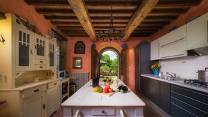 A kitchen with a central island, wooden beams, and a view to an outdoor patio. - Podere Santa Croce 10, Emma Villas (Radicondoli)