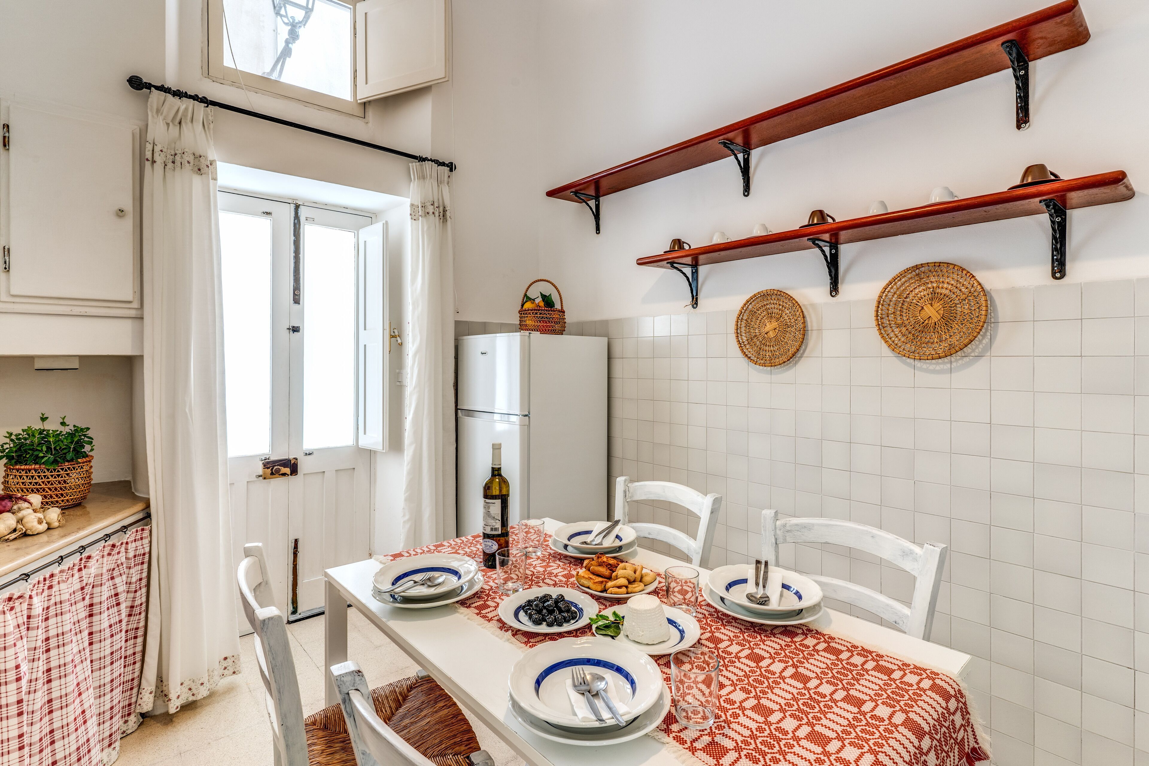 Comedor con una mesa lista para cuatro, sillas blancas, mantel rojo y blanco con estampado, y un salpicadero de azulejos blancos.