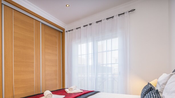 Habitación de hotel con un armario de madera, una cama con colcha roja y negra, y una ventana con cortinas translúcidas.