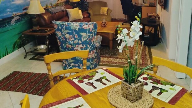 A living room with a mural of a tropical scene, a chandelier, a sofa, a coffee table, and a dining area with a table set for two. - Unique 2 BR, 2 Bath Condo in Surfside Beach, SC;  Central Grand Strand (Surfside Beach)