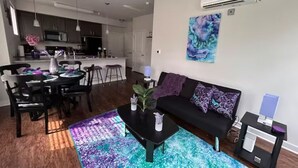 Een moderne woonkamer met een zwarte bank, een salontafel en een eethoek met een tafel en stoelen. Achtergrond is een keuken met kasten en een magnetron.