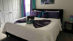 Een net opgemaakt bed met een witte deken, paarse en teal kussens, en een mand met handdoeken. Daarboven hangen drie schilderijen aan de muur.
