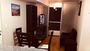 Een compact woongedeelte met een keukenhoek, voorzien van een granieten aanrechtblad en houten kastjes. Er staat een eettafel met stoelen, een zwarte bank en een televisie op de achtergrond.