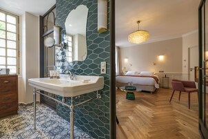 Ένα μπάνιο με νιπτήρα, καθρέφτη και διακοσμητικά πλακάκια. Το δωμάτιο διαθέτει ξύλινο δάπεδο και ένα κρεβάτι με λευκά σεντόνια στο βάθος.