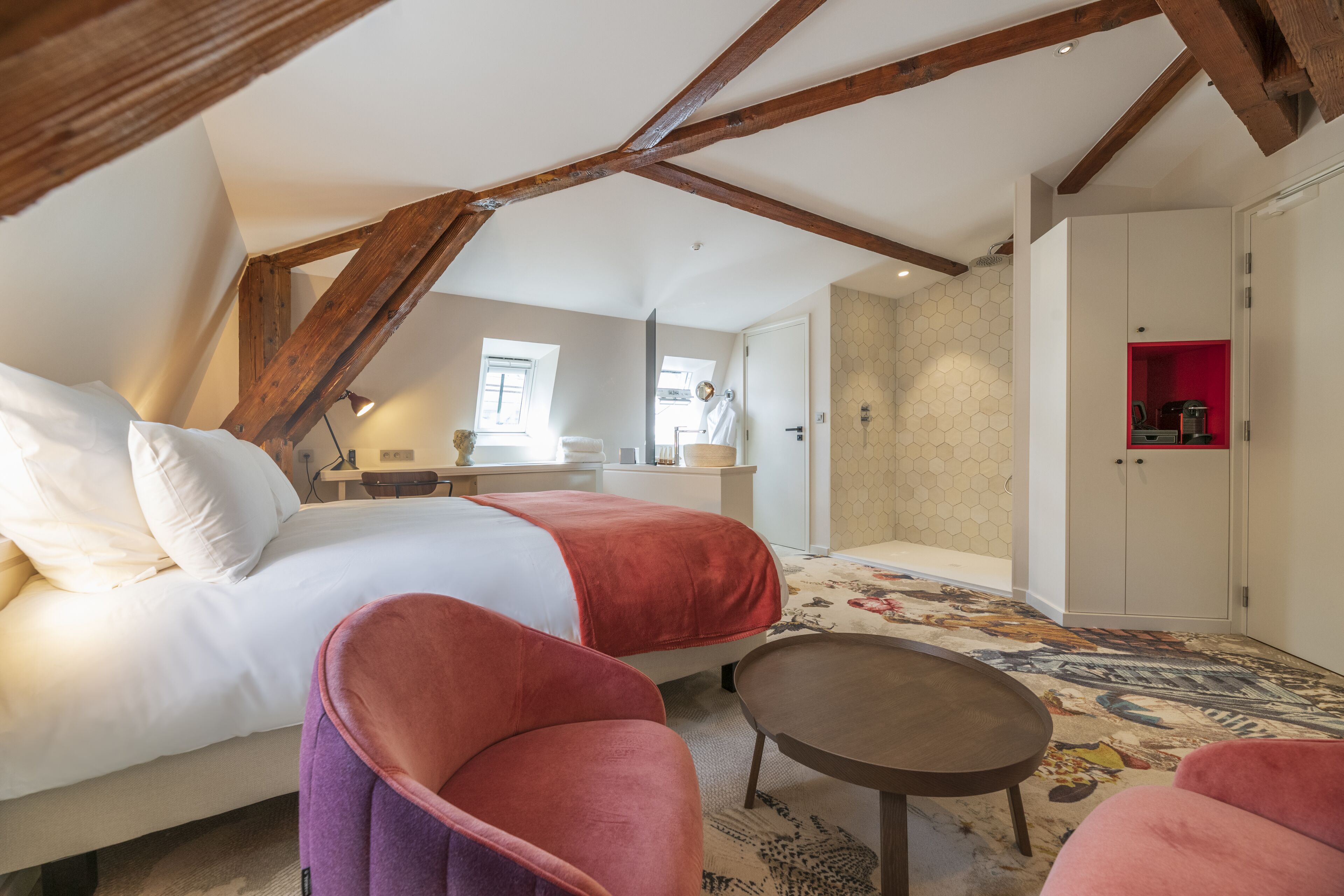 A bedroom with a bed, two pink chairs, a small table, and a bathroom with a shower and toilet.