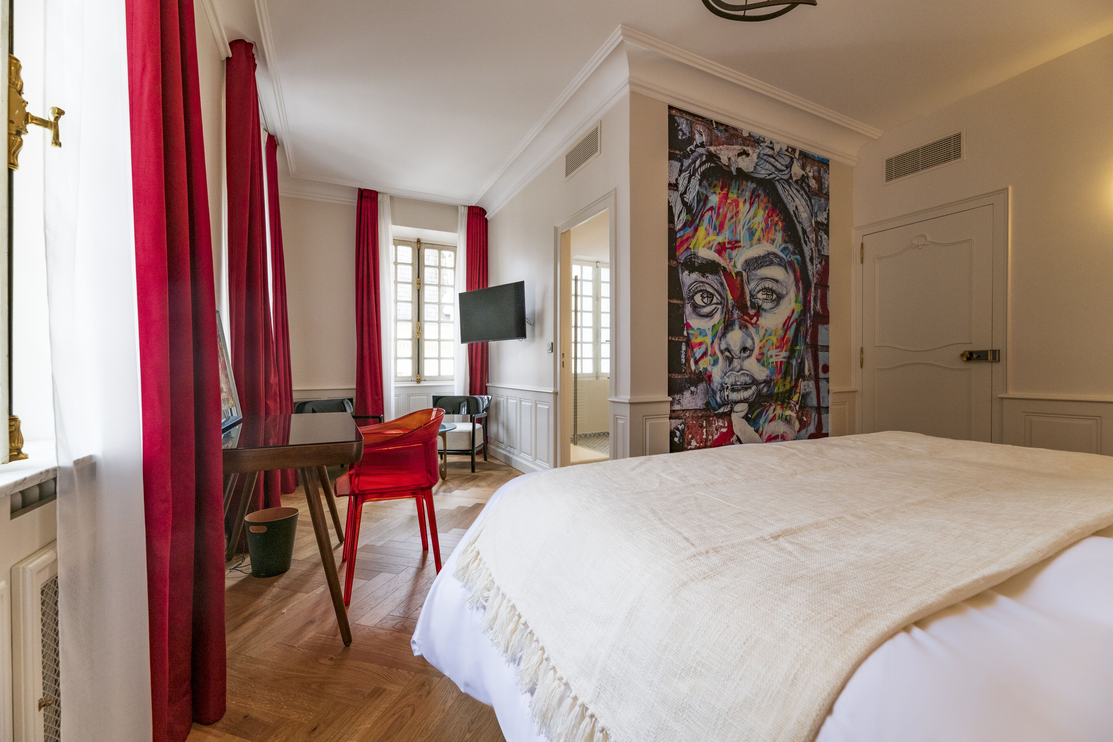 Ein Hotelzimmer mit einem großen Bett, einem Schreibtisch mit Stuhl, einem Fernseher und einem farbenfrohen Wandbild.