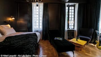 Ett hotellrum med mörkt tema, utrustat med en stor säng, en läsbänk, ett litet bord med glas och en flaska samt en stol.