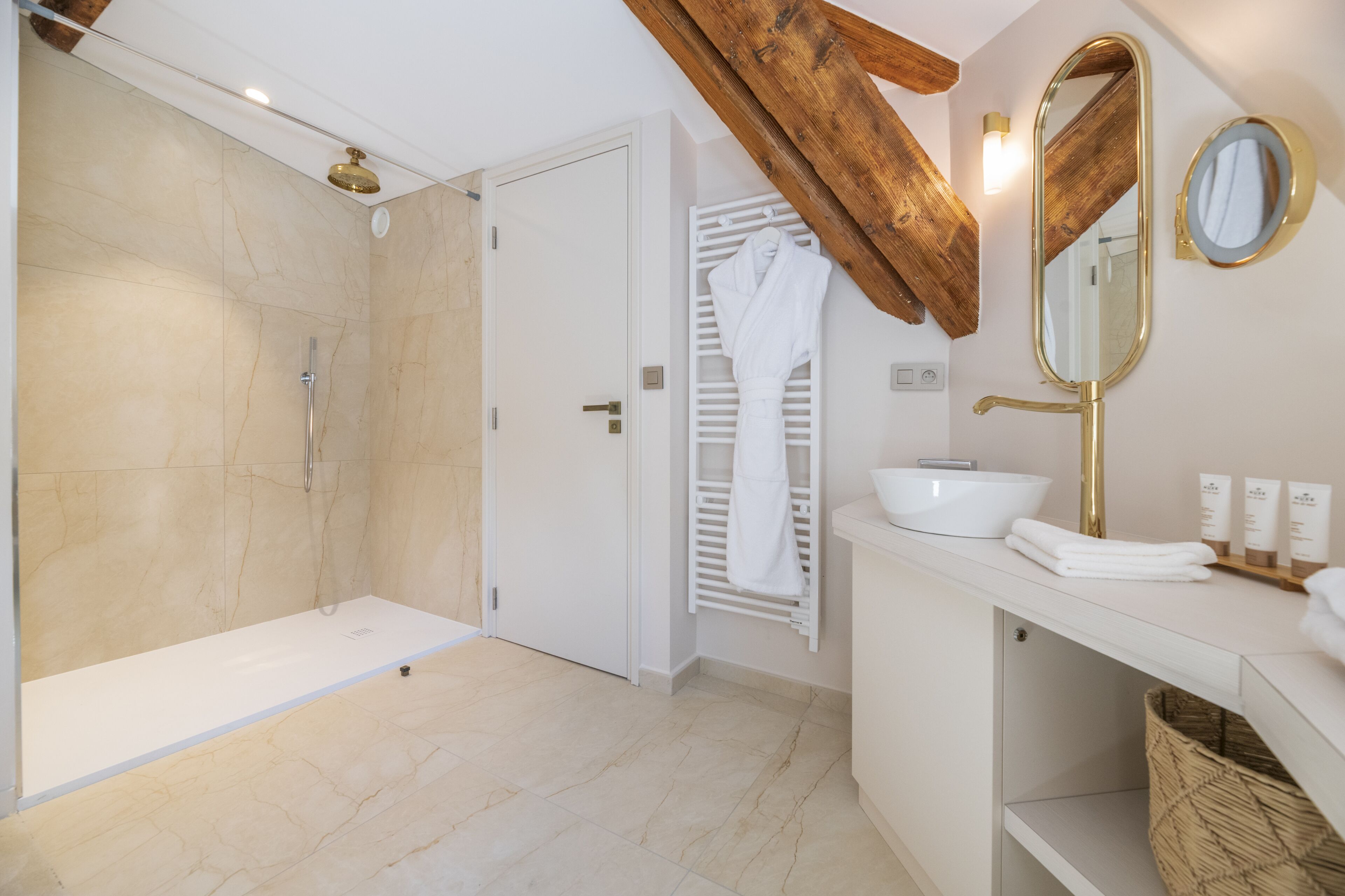 Ein modernes Badezimmer mit bodenebenen Dusche, einem weißen Waschtisch mit goldfarbenem Wasserhahn und einem Holzbalken an der Decke.