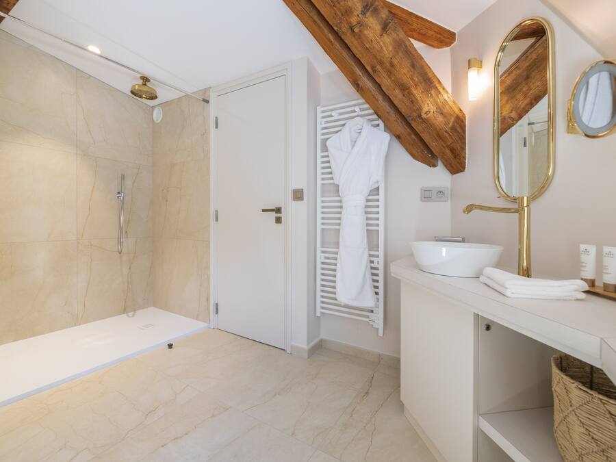 Ein modernes Badezimmer mit bodenebenen Dusche, einem weißen Waschtisch mit goldfarbenem Wasserhahn und einem Holzbalken an der Decke.