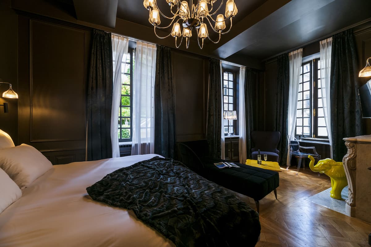 A large bed with a dark throw, a desk with a chair, and a yellow elephant sculpture in a room with dark walls and large windows.