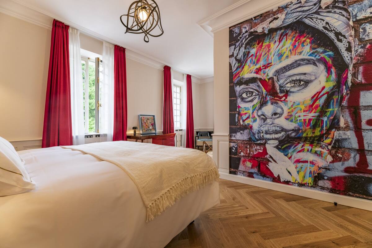 A hotel room with a large bed, red curtains, a painting on the wall, and a wooden floor.