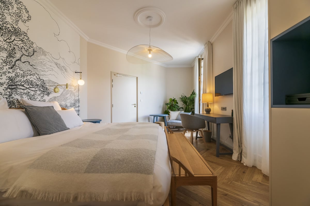 A modern hotel room with a large bed, a desk, a chair, a small table, a wall-mounted TV, and a large window with curtains.