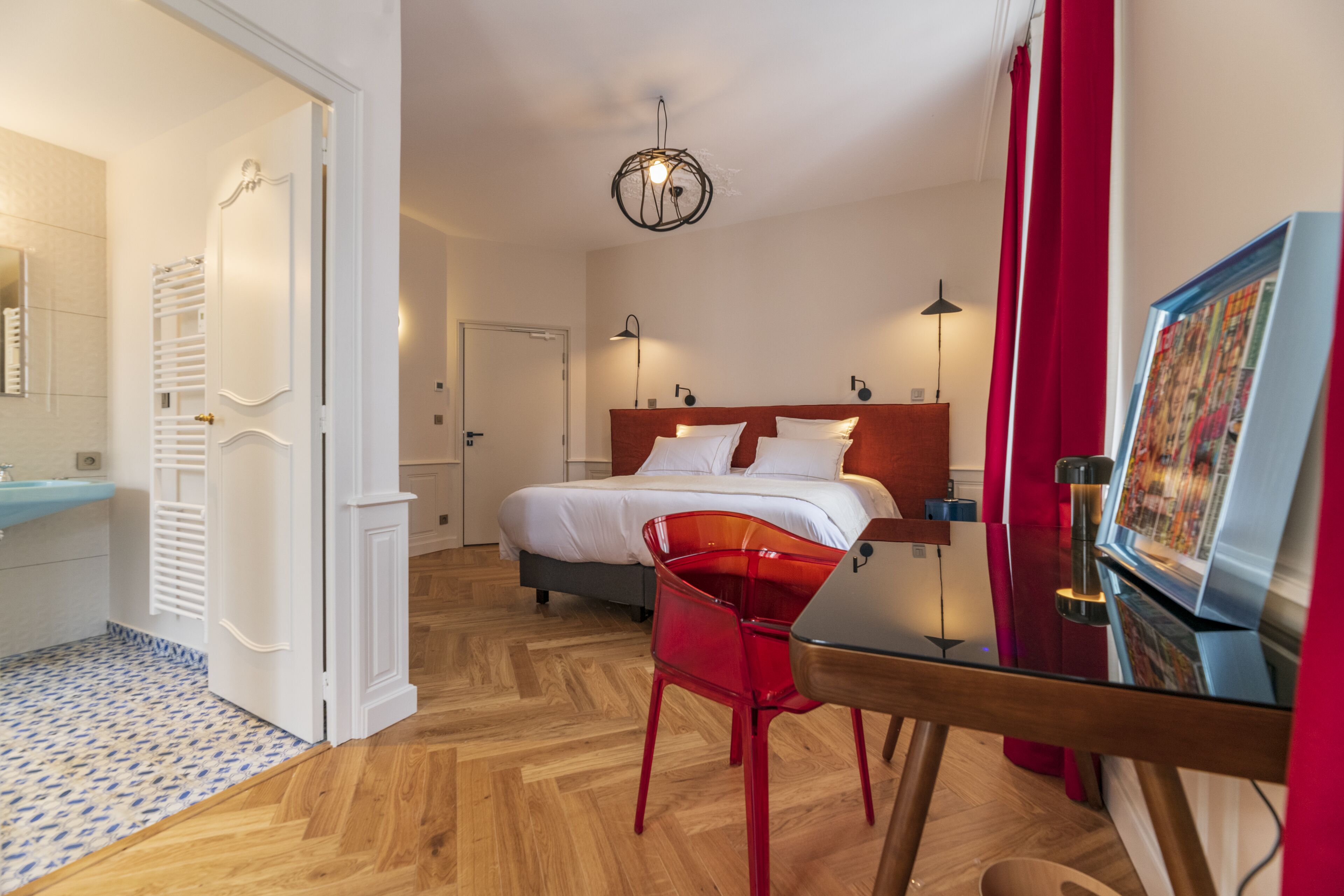 Une chambre d’hôtel avec un lit, une chaise rouge, un bureau, une télévision et une salle de bain équipée d’un miroir et d’un porte-serviettes.