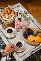 Una bandeja con alimentos para el desayuno, incluyendo cruasanes, un bol de cerezas, un kiwi, una naranja y una taza de café.