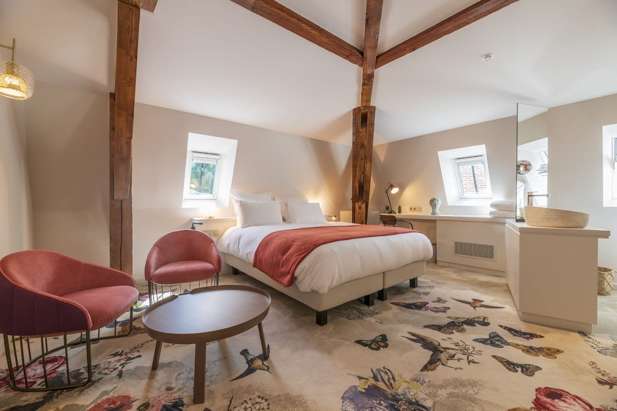 A bedroom with a bed, two red chairs, a round table, and a large mirror.