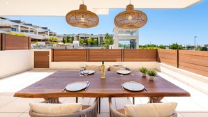 A dining area with a wooden table set for four, two hanging wicker lamps, and a view of modern buildings in the background. - Jardiana Lotus | Walking distance to La Cala De Mijas (La Cala de Mijas)