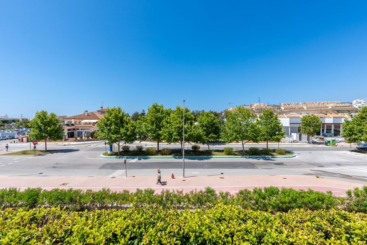 A wide street with a pedestrian crossing, trees, and buildings in the background.