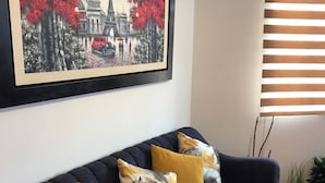 A dark blue sofa with three cushions in different colors, a framed picture of a cityscape on the wall, and a window with horizontal blinds.