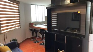 A modern kitchen with a large black TV, a dining area with bar stools, and a blue sofa in a living room area.