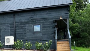 A modern building with a dark exterior, a small window, and a covered porch with a wooden staircase. - Private Driftless Cabin w/ Hot Tub, Fireplace & Creekside Views (Ferryville)