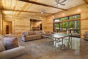 A spacious room with a ping pong table, a sofa, and a large window offering a view of the forest.