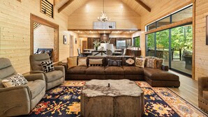 A spacious living room with a large sectional sofa, a coffee table, and a fireplace. There is a dining area with a table and chairs, and a kitchen area with a refrigerator and stove. The room has large windows and a wooden ceiling. - Bison's Bend in Hochatown/Broken Bow, OK (Broken Bow)