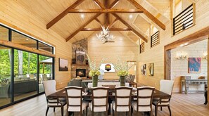 A spacious wooden interior with a high ceiling, a fireplace, dining area, and large windows overlooking a forested area.