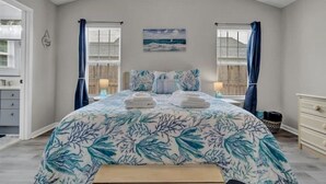 Un dormitorio con cama, dos mesitas de noche, una mesita auxiliar, una ventana con cortinas, una obra de arte en la pared y vistas al exterior.
