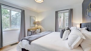 A hotel room with a large bed, a desk, and a chair. There are windows with curtains, a lamp, and a wall-mounted light fixture. - Hot Tub, Fire Pit, Ping-Pong, 3 min walk to Olde Town Arvada! (Arvada)
