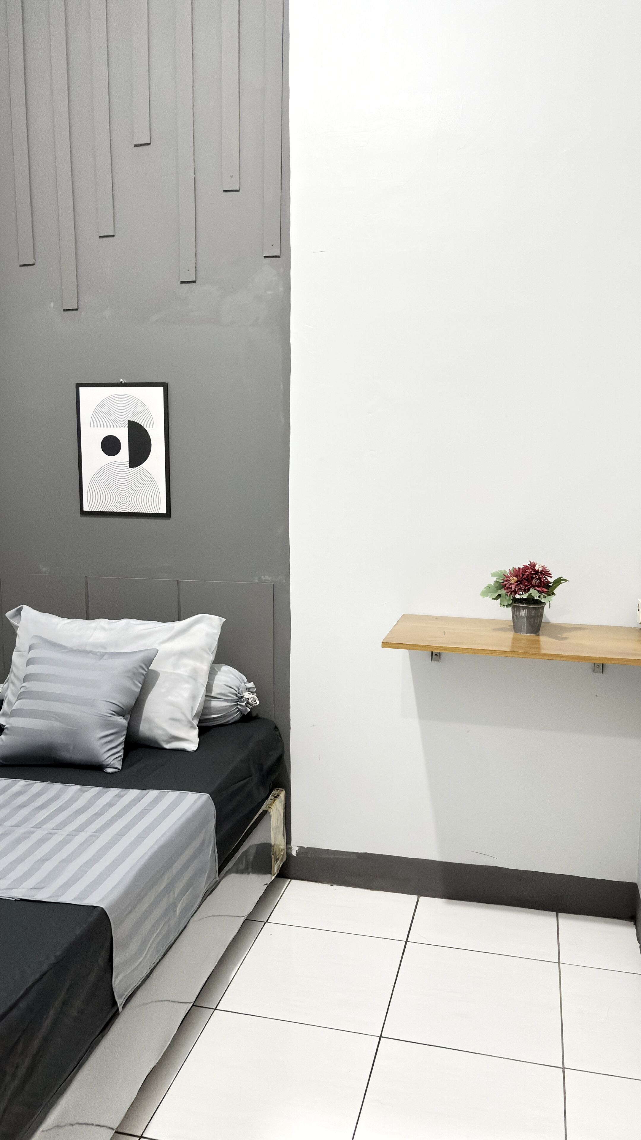 Un dormitorio con una cama, almohadas, un cuadro enmarcado, un estante pequeño con una planta y suelo de baldosas.