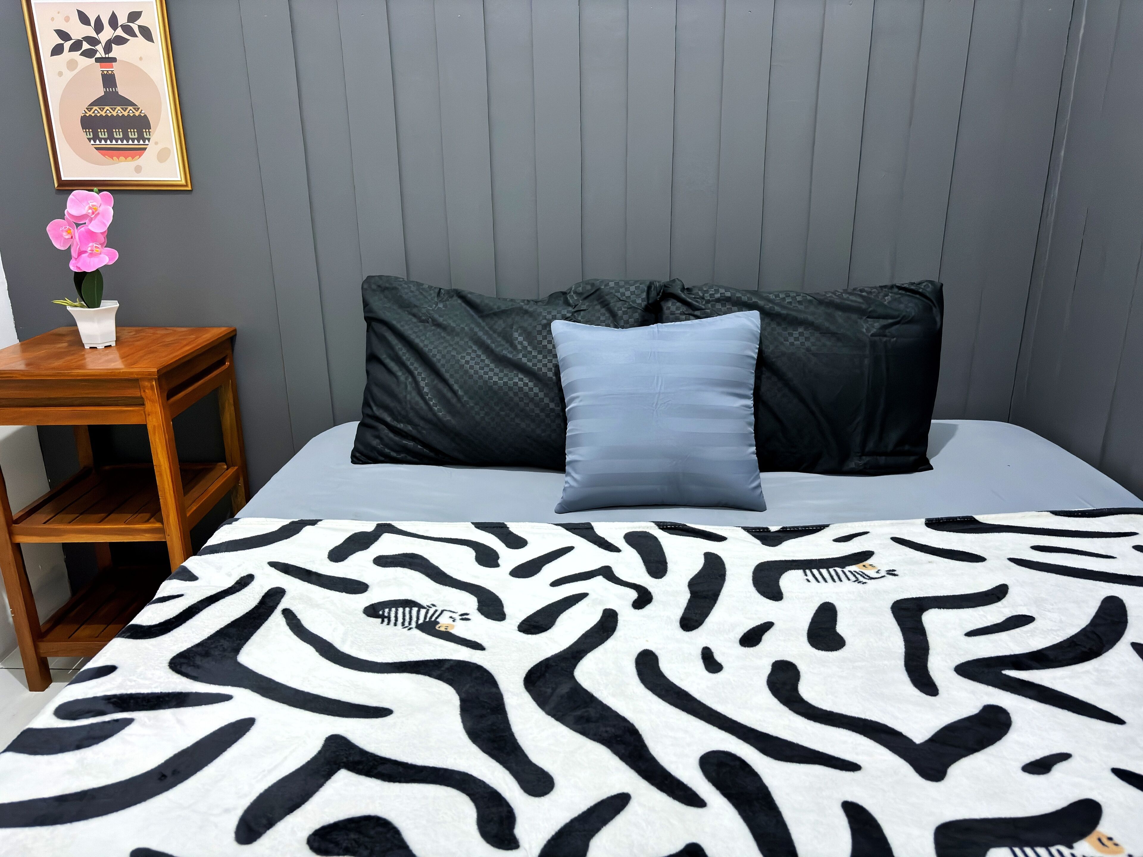 A bedroom with a zebra-patterned bedspread, a grey wall, a framed picture, a small wooden nightstand, and a pink orchid plant.