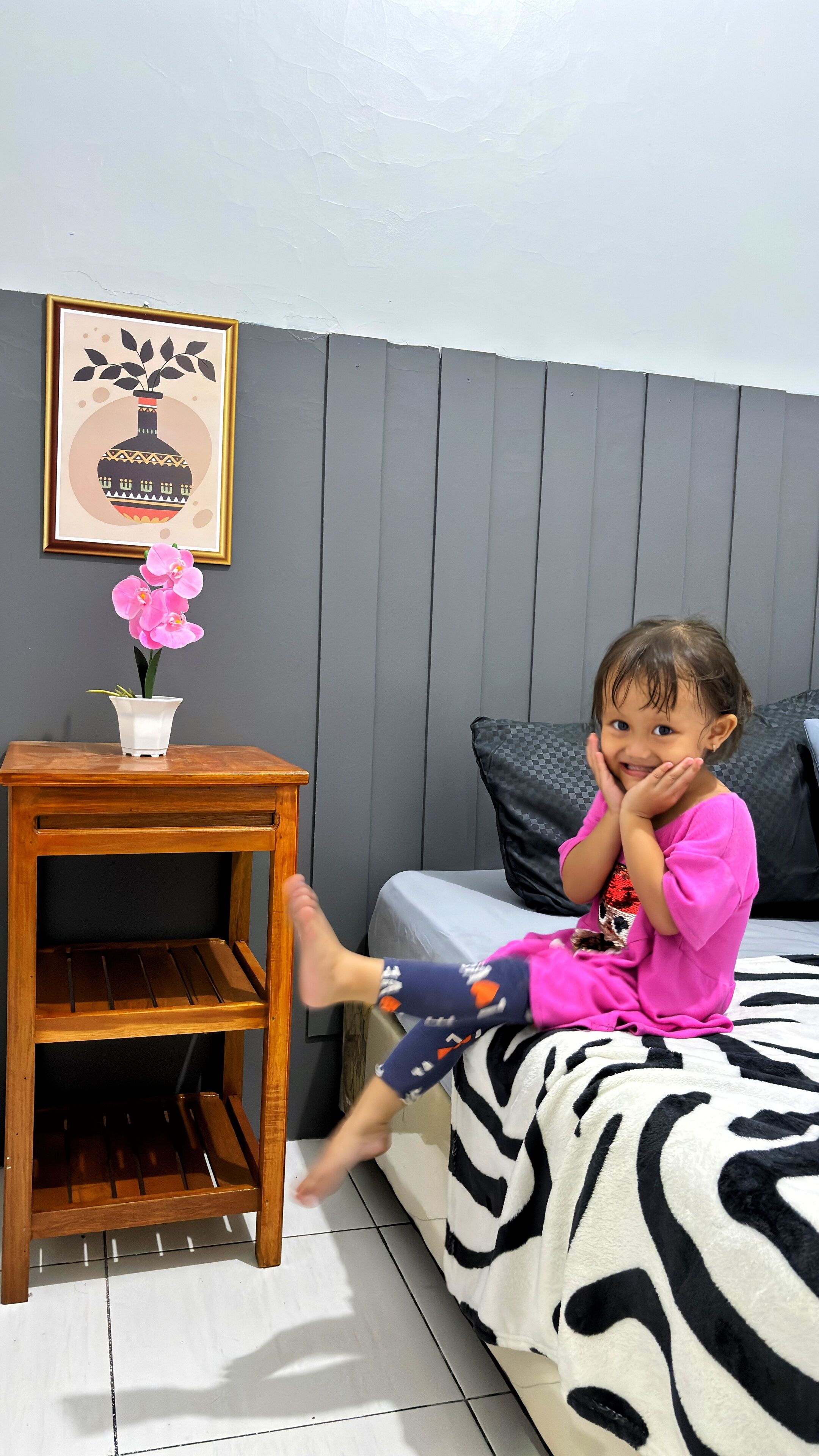 Un niño sentado en una cama con una manta de estampado de cebra, una mesita de noche de madera con un jarrón blanco y una orquídea rosa, y un cuadro en la pared.