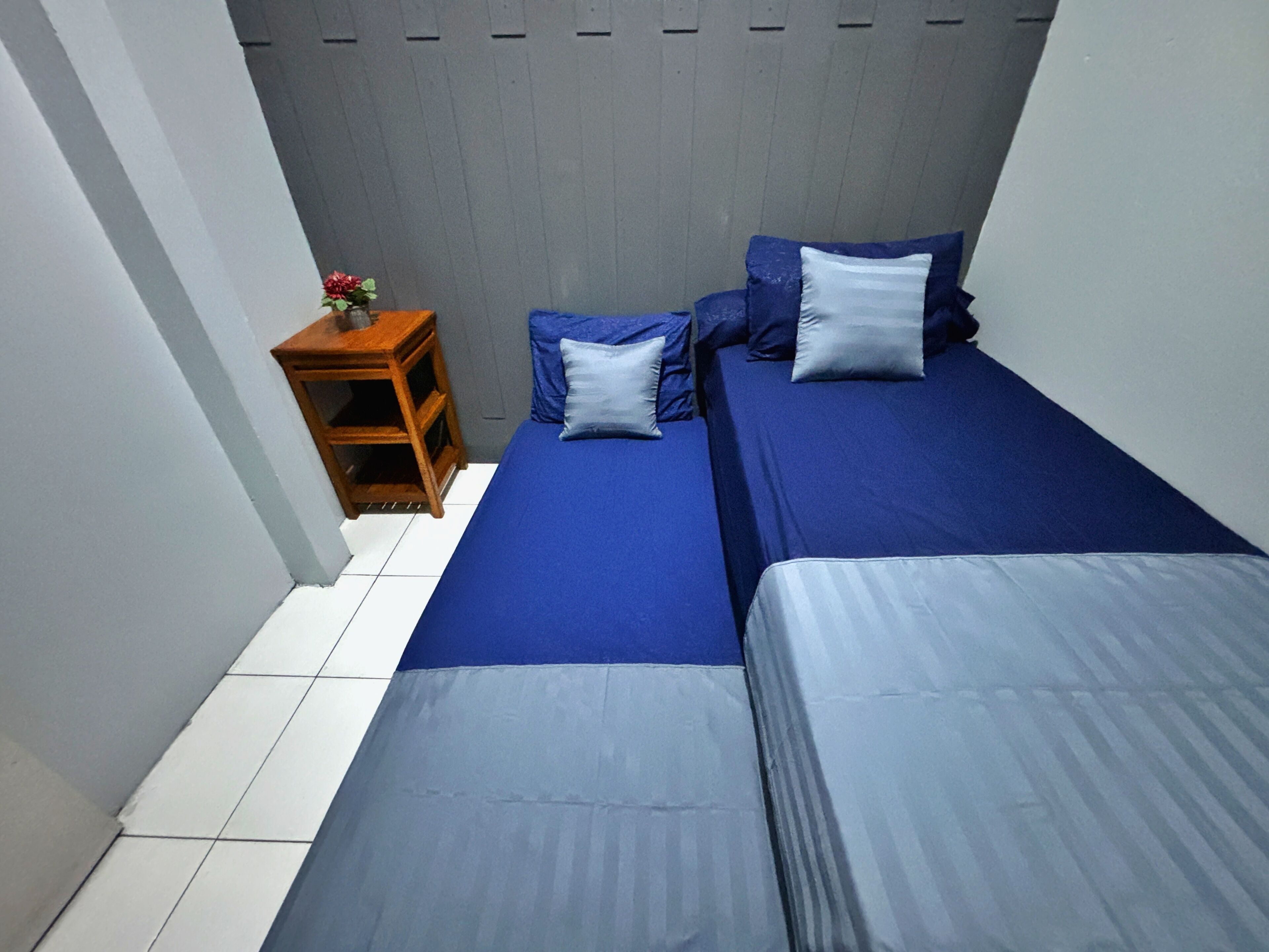 A small, narrow room with two beds, a nightstand, and a small table with a vase of flowers.