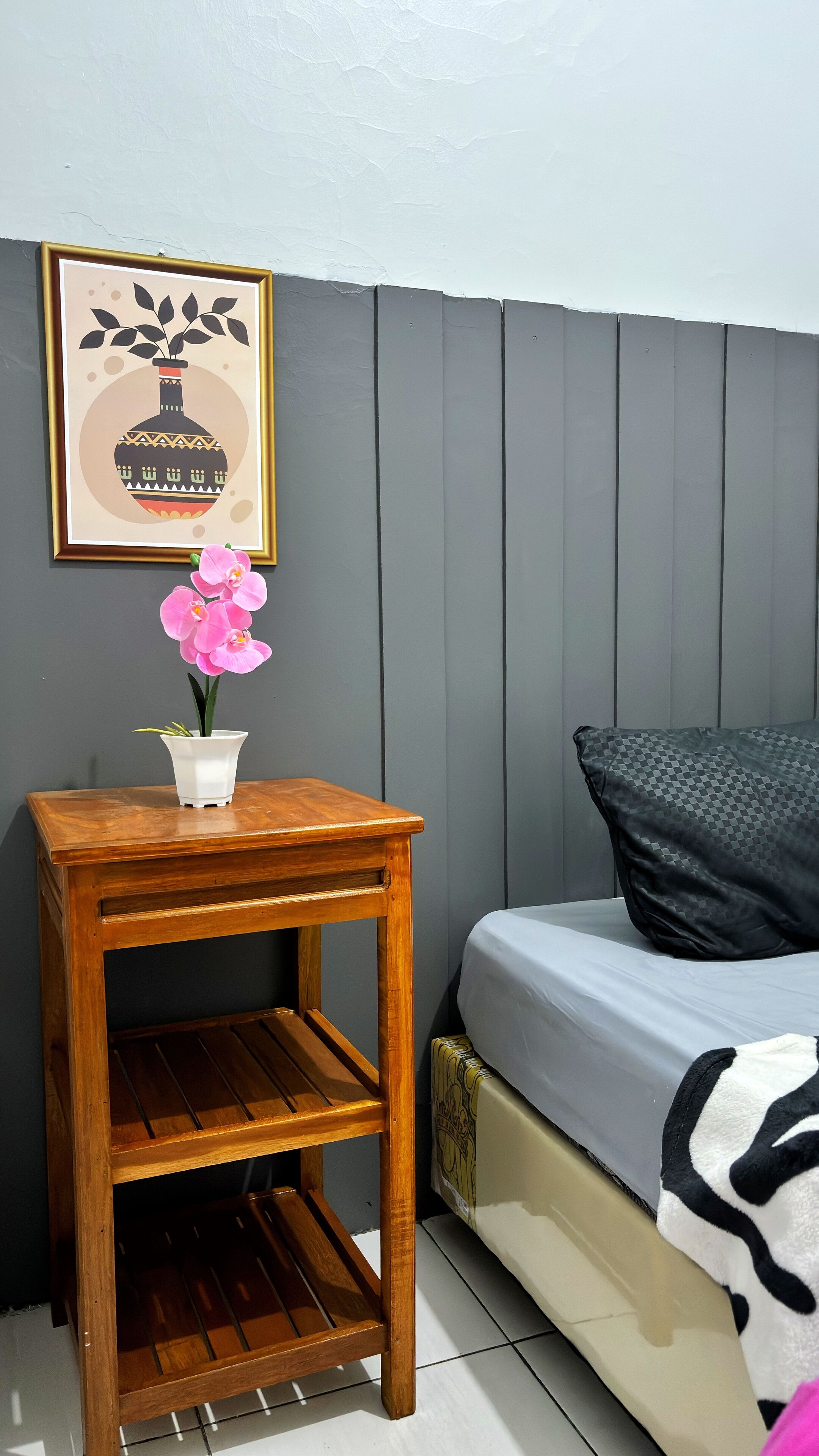 A small wooden nightstand with a white vase and pink orchid, a framed picture above it, and a bed with a checkered blanket and black pillow against a grey wall.