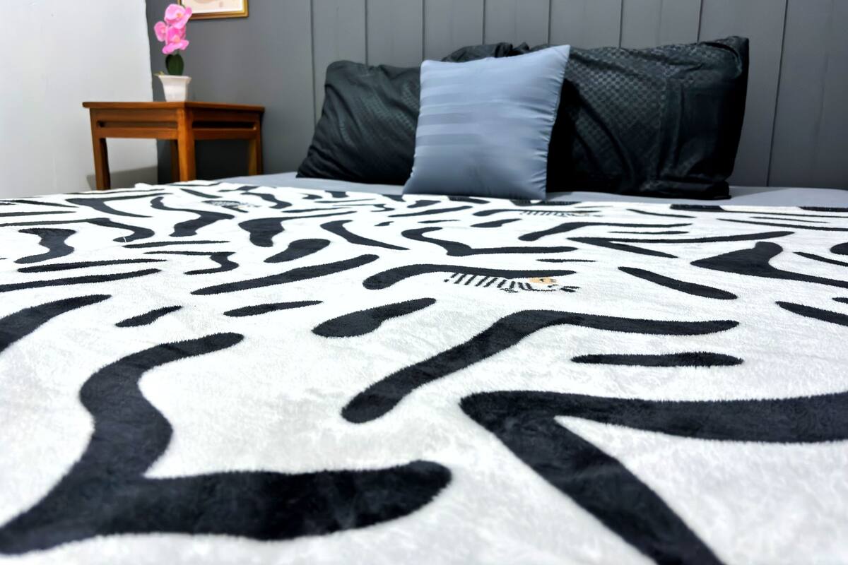 A bed with a zebra print blanket, a small wooden nightstand with a vase of flowers, and a framed picture on the wall.