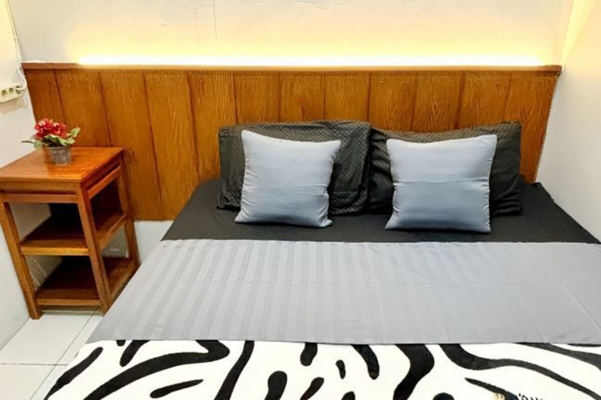 A bedroom with a wooden headboard, two pillows, a small wooden nightstand with a flower vase, and a zebra-patterned rug.