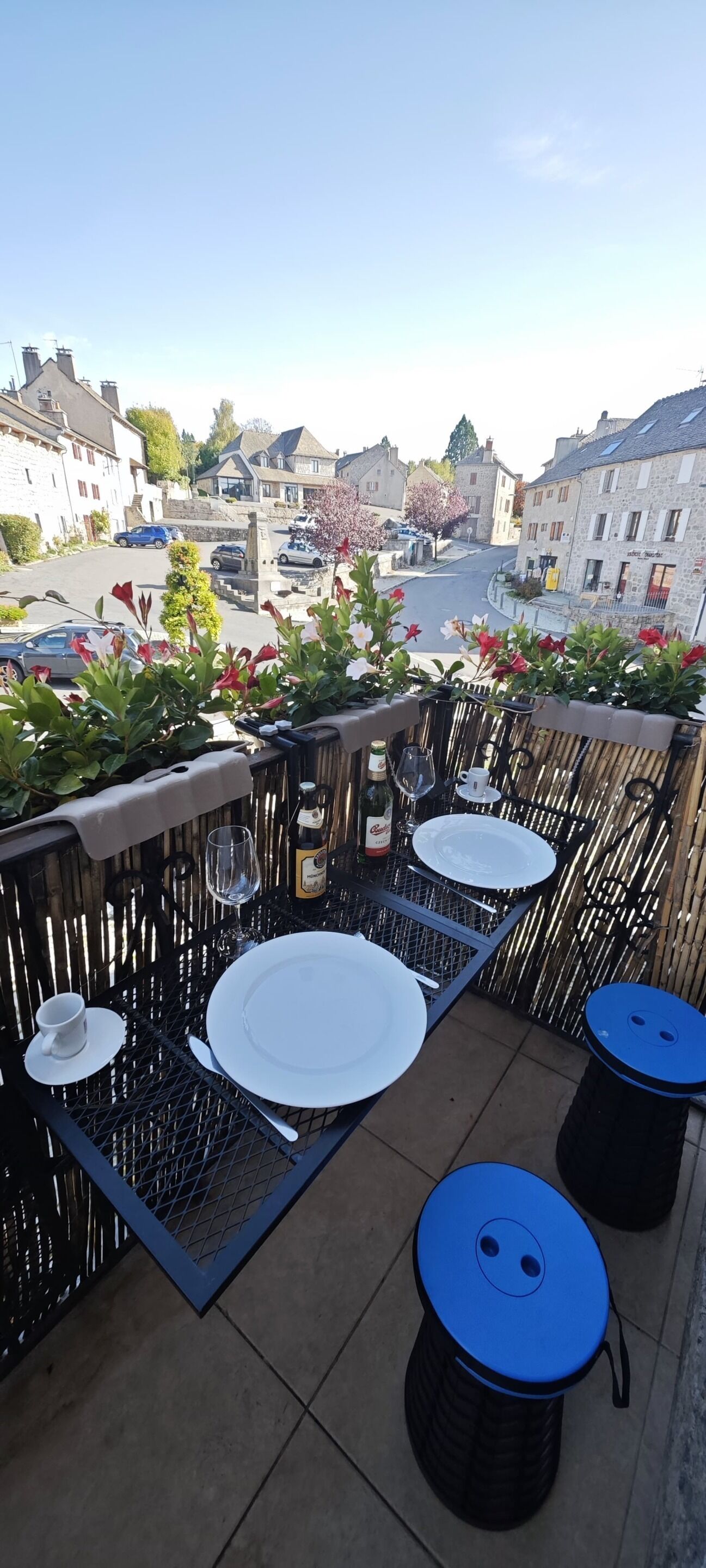 Un balcon avec une table dressée pour deux personnes, une bouteille de bière et un verre. On y trouve des plantes en pot et une vue sur une rue bordée d’immeubles et de voitures.