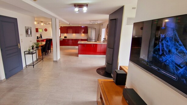 Un salon moderne avec un téléviseur à écran plat, un coin repas avec une cuisine rouge en arrière-plan, et un couloir avec une porte et un radiateur.