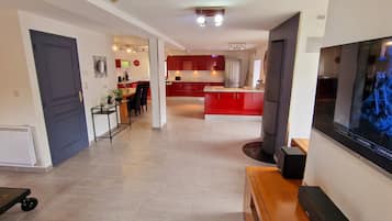 Un salon moderne avec un téléviseur à écran plat, un coin repas avec une cuisine rouge en arrière-plan, et un couloir avec une porte et un radiateur.