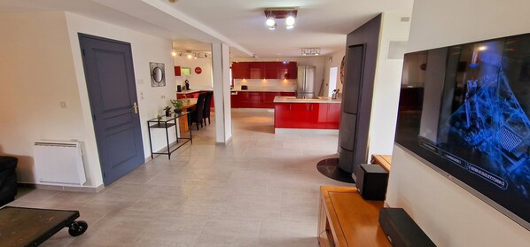 A modern living room with a flat-screen TV, a dining area with a red kitchen in the background, and a hallway with a door and radiator. - Gîte le Fortunio-Gîte-Family-Ensuite with Bath (RIEUTORT DE RANDON)