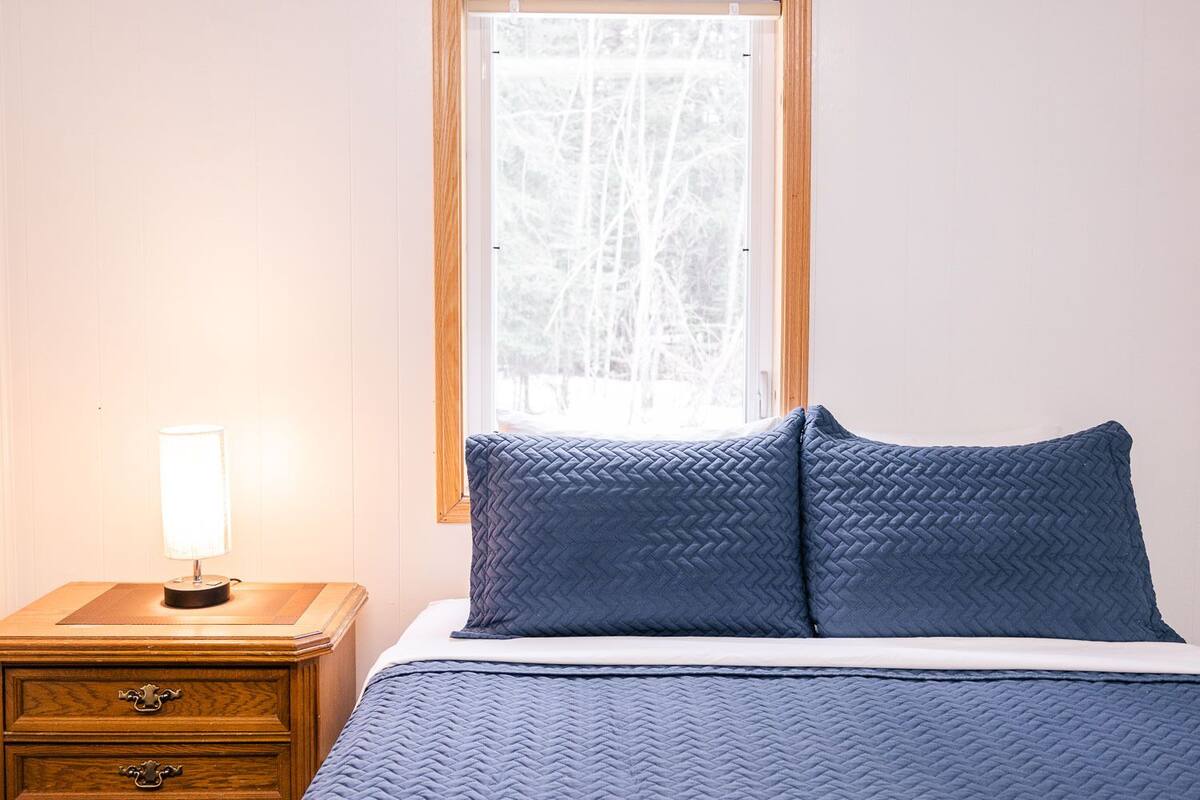 Un dormitorio con cama, mesita de noche con lámpara y ventana con vista a árboles.