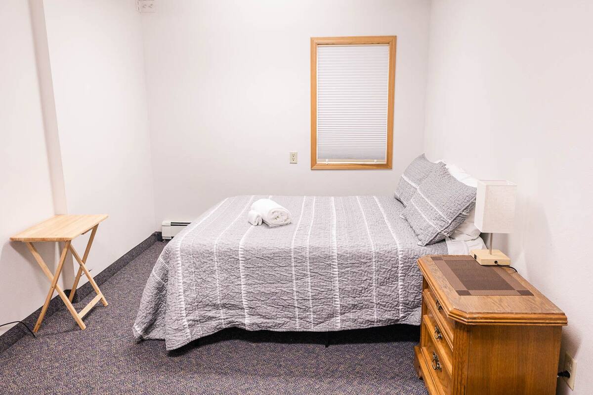 Un dormitorio con una cama, una mesita de noche de madera, una pequeña mesa de madera y una ventana con persianas.