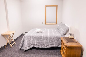 A bedroom with a bed, a wooden nightstand, a small wooden table, and a window with blinds.