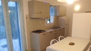 Une cuisine compacte équipée d’une cuisinière à gaz, d’un évier et de placards, d’une table à manger avec des chaises, et d’une grande fenêtre avec des rideaux.